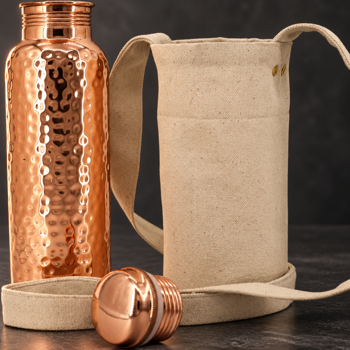 Close-up of an open hammered copper-colored bottle, matching screw cap, and beige shoulder bag with a long strap.