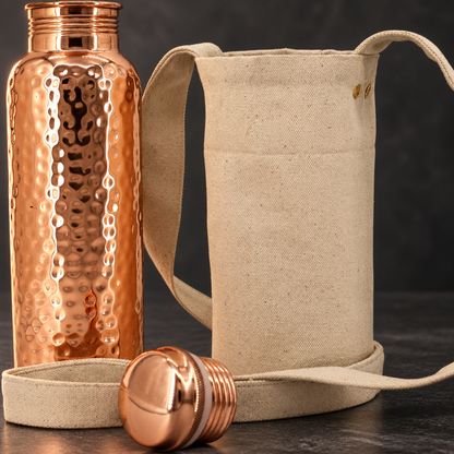 Close-up of an open hammered copper-colored bottle, matching screw cap, and beige shoulder bag with a long strap.